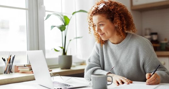 Cute university student with curly red hair doing homework sitting at kitchen table with coffee cup and wired earphones next to big window, watching online tutorial on laptop and writing in copybook