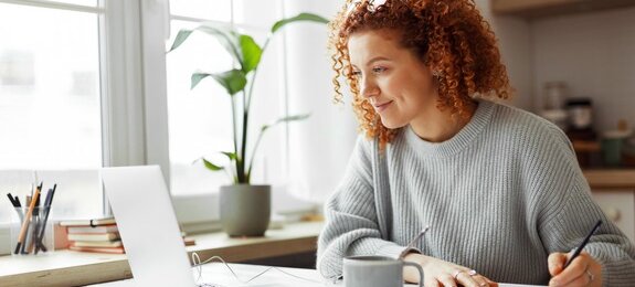 Cute university student with curly red hair doing homework sitting at kitchen table with coffee cup and wired earphones next to big window, watching online tutorial on laptop and writing in copybook