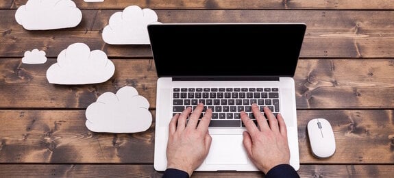 Cloud computing technology concept with white fluffy clouds next to the laptop. Mans hands typing the the keyboard uploading data, on a wooden table.