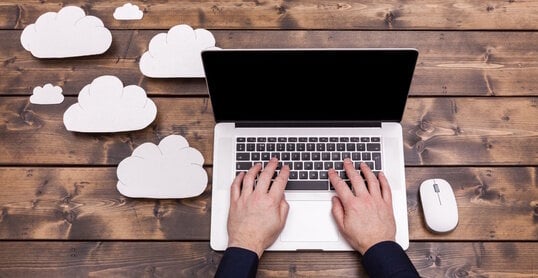 Cloud computing technology concept with white fluffy clouds next to the laptop. Mans hands typing the the keyboard uploading data, on a wooden table.