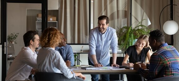 Happy male business team leader talking to employees at corporate meeting, discussing work project, sharing jokes, laughing. Positive mentor training diverse group of interns, having fun