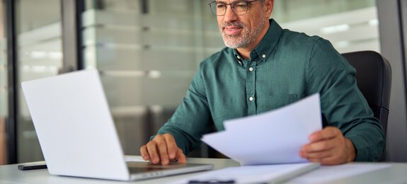 Smiling middle aged executive, mature male hr manager holding documents using laptop looking at pc computer in office at desk, checking financial data in report, doing account paper plan overview.