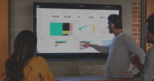 Drei Personen stehen vor einem Bildschirm und unterhalten sich über Diagramme, die dort abgebildet sind. Die Diagramme sind dabei in dem CRM-System Microsoft Dynamics 365 sichtbar.