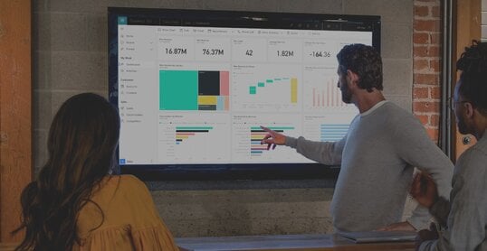 Drei Personen stehen vor einem Bildschirm und unterhalten sich über Diagramme, die dort abgebildet sind. Die Diagramme sind dabei in dem CRM-System Microsoft Dynamics 365 sichtbar.