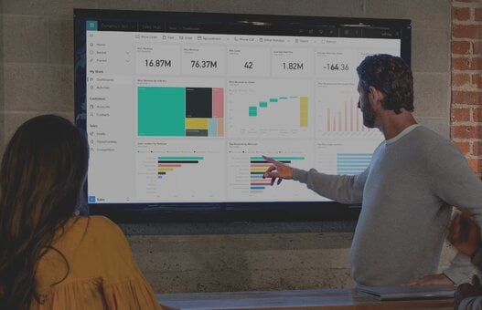 Drei Personen stehen vor einem Bildschirm und unterhalten sich über Diagramme, die dort abgebildet sind. Die Diagramme sind dabei in dem CRM-System Microsoft Dynamics 365 sichtbar.