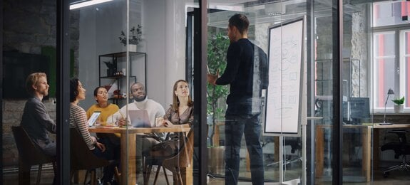 Project Manager Makes a Presentation for a Young Diverse Creative Team in Meeting Room in an Agency. Colleagues Sit Behind Conference Table and Discuss Business Development, User Interface and Design. 