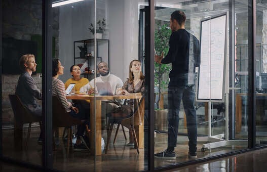 Project Manager Makes a Presentation for a Young Diverse Creative Team in Meeting Room in an Agency. Colleagues Sit Behind Conference Table and Discuss Business Development, User Interface and Design. 