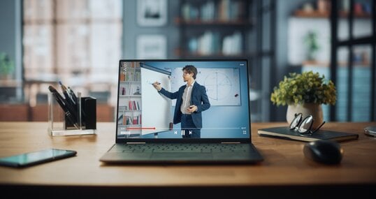 Shot of a Laptop Computer Showing Online Lecture with Portrait of a Cute Male Teacher Explaining Math Formulas. It is Standing on the Wooden Desk in Stylish Modern Home Office Studio During Day.