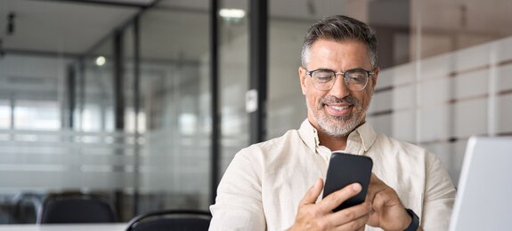 Middle aged Hispanic business manager ceo using cell phone mobile app, laptop. Smiling Latin or Indian mature man businessman holding smartphone sit in office working online on gadget with copy space.