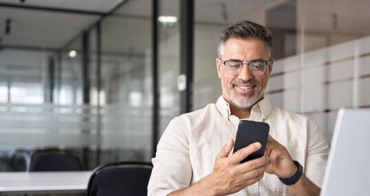 Middle aged Hispanic business manager ceo using cell phone mobile app, laptop. Smiling Latin or Indian mature man businessman holding smartphone sit in office working online on gadget with copy space.