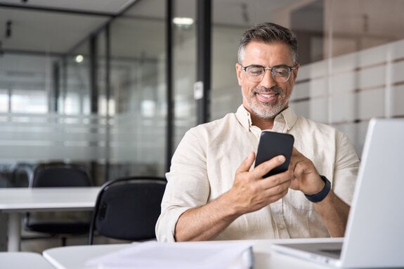 Middle aged Hispanic business manager ceo using cell phone mobile app, laptop. Smiling Latin or Indian mature man businessman holding smartphone sit in office working online on gadget with copy space.