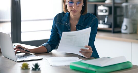 Young business woman working with computer while consulting some invoices and documents in the kitchen at home.