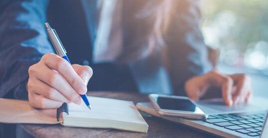 Business woman hand writing on a notepad with a pen and using laptop in the office.