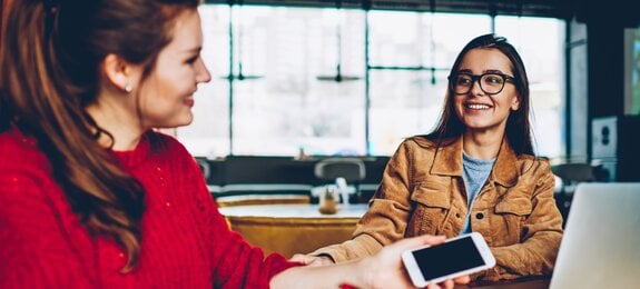 Two happy female friends enjoying positive conversation looking to each other in break of learning and searching information for test indoors, smiling hipster girls talking and spending time together