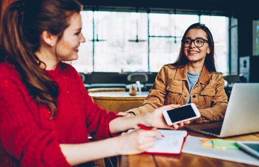 Two happy female friends enjoying positive conversation looking to each other in break of learning and searching information for test indoors, smiling hipster girls talking and spending time together