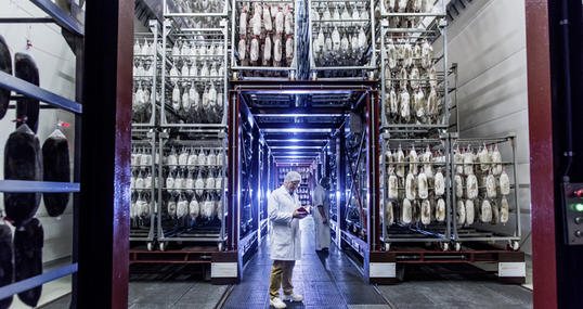Foto einer Fabrikhalle der Bell Food Group mit zwei Mitarbeitern in Schutzkleidung und Waren die von Regalen hängen